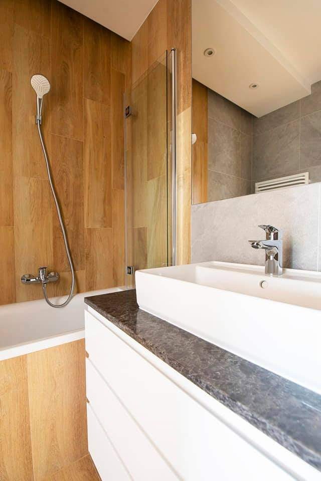 Nowoczesna łazienka z wanną obłożoną drewnopodobnymi płytkami, szklanym parawanem nawannowym, białą umywalką nablatową na blacie z ciemnego kamienia i szarą płytką na ścianie z lustrem.