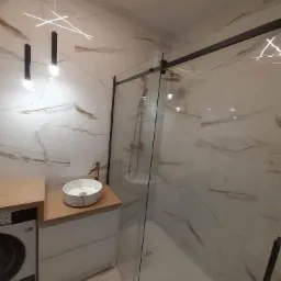 Nowoczesna łazienka z kabiną prysznicową typu walk-in, umywalką nablatową na drewnianym blacie, marmurowymi płytkami ściennymi z delikatnymi żyłami i designerską lampą wiszącą.