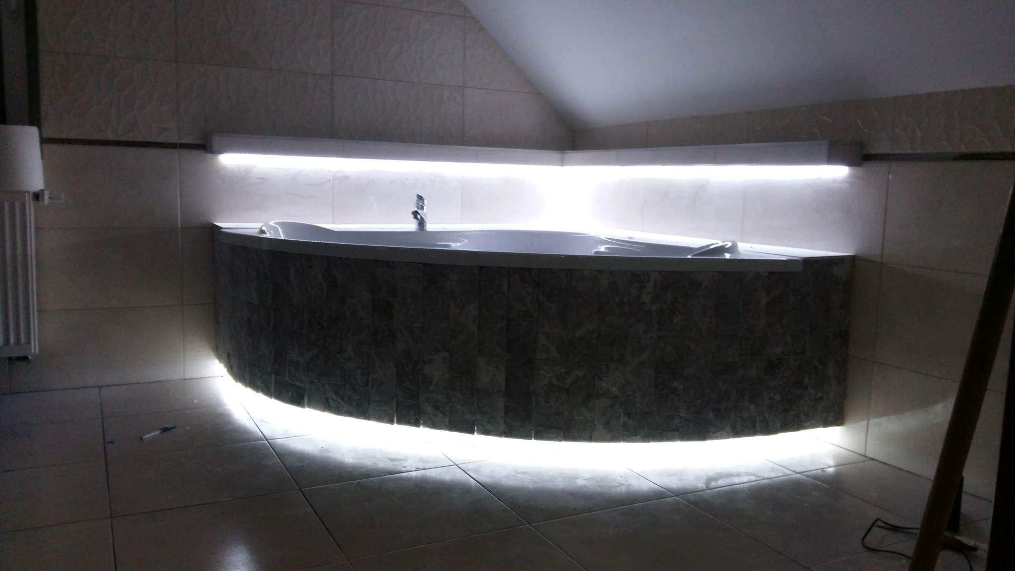 Narożna wanna z obudową z ciemnych płytek imitujących kamień, podświetlona taśmą LED od dołu i od góry, umieszczona w łazience z jasnymi, kwadratowymi płytkami na ścianach i podłodze.