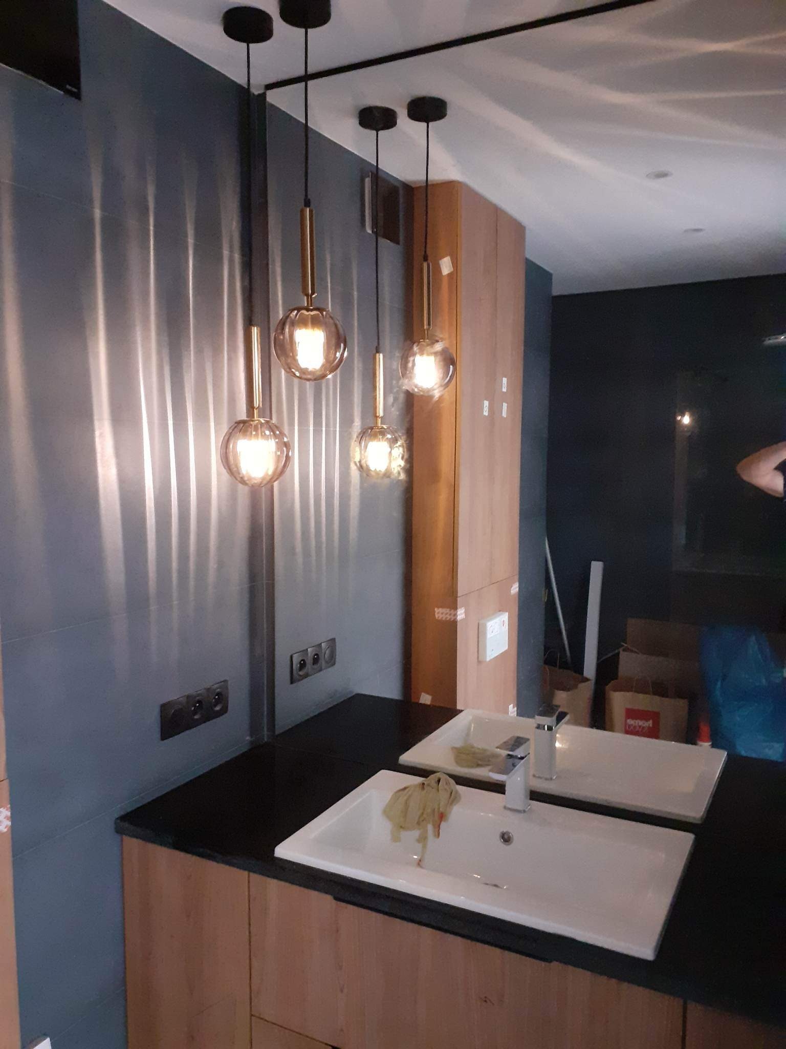 Nowoczesna łazienka z trzema szklanymi lampami wiszącymi na czarnych przewodach, odbijającymi się w lustrze nad umywalkami z czarnym blatem, szare płytki na ścianie, drewniane szafki pod umywalkami...