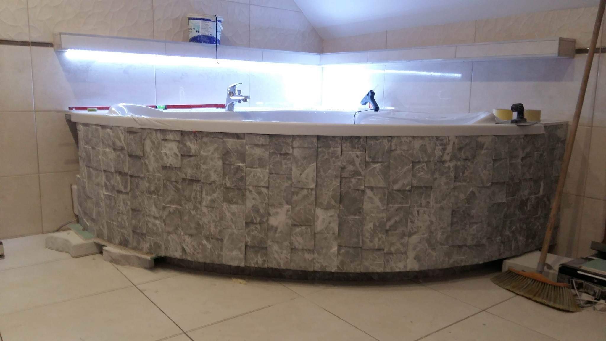 Obudowa wanny z mozaiki kamiennej w trakcie montażu, widoczne poziomica, pistolet do kleju i wiaderko z klejem, oświetlenie LED pod półką.