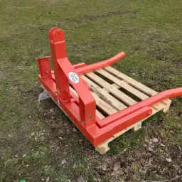 Czerwony, metalowy osprzęt do traktora - widły do palet z zamontowaną paletą, stojący na trawie.