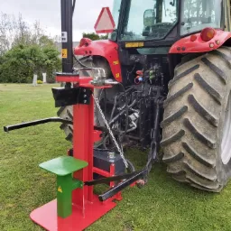 Czerwono-czarna łuparka do drewna z zielonym stołem roboczym, podłączona do czerwonego ciągnika rolniczego na trawniku.