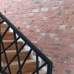 Klatka schodowa z drewnianymi stopniami, czarną metalową balustradą o geometrycznym wzorze i ścianą wyłożoną cegłą klinkierową w odcieniach czerwieni i brązu. Widoczne fragmenty białej listwy...