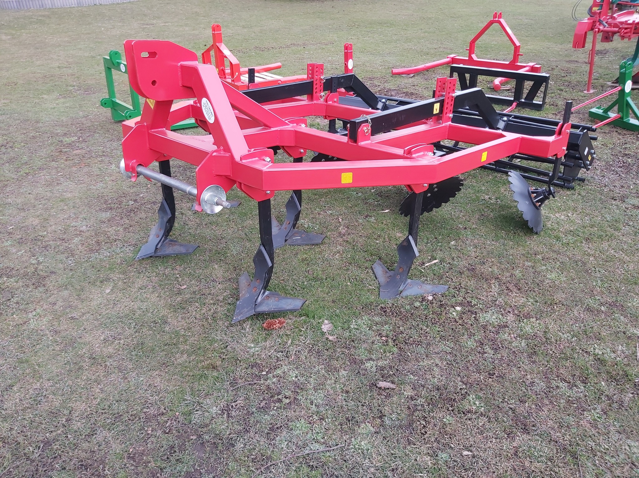 Czerwony kultywator z czarnymi elementami roboczymi, stojący na trawie z innymi maszynami rolniczymi w tle.