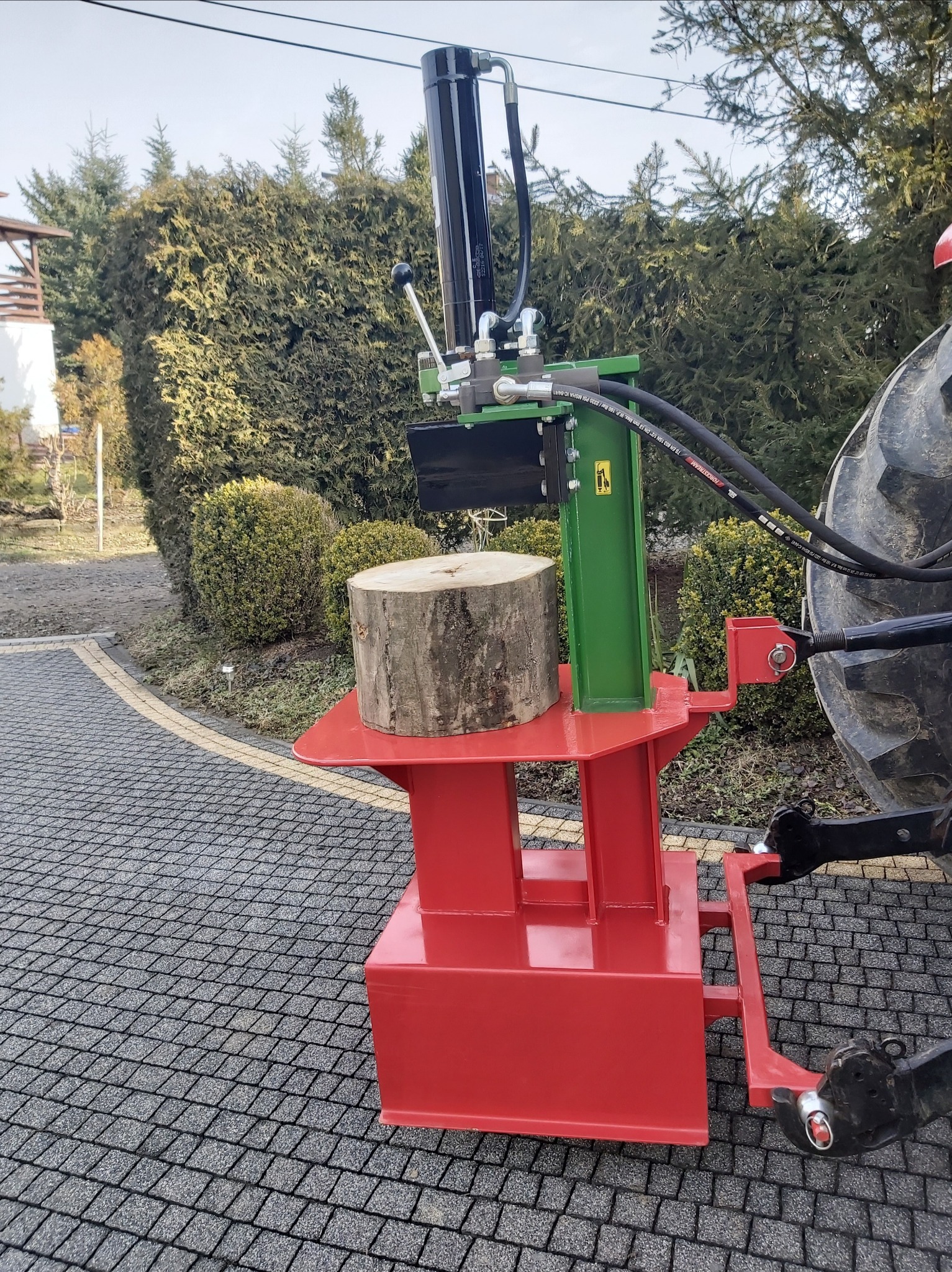 Hydrauliczna łuparka do drewna, pomalowana na czerwono i zielono, zamontowana na traktorze, z kłodą drewna przygotowaną do rozłupania, tło stanowią krzewy i drzewa.