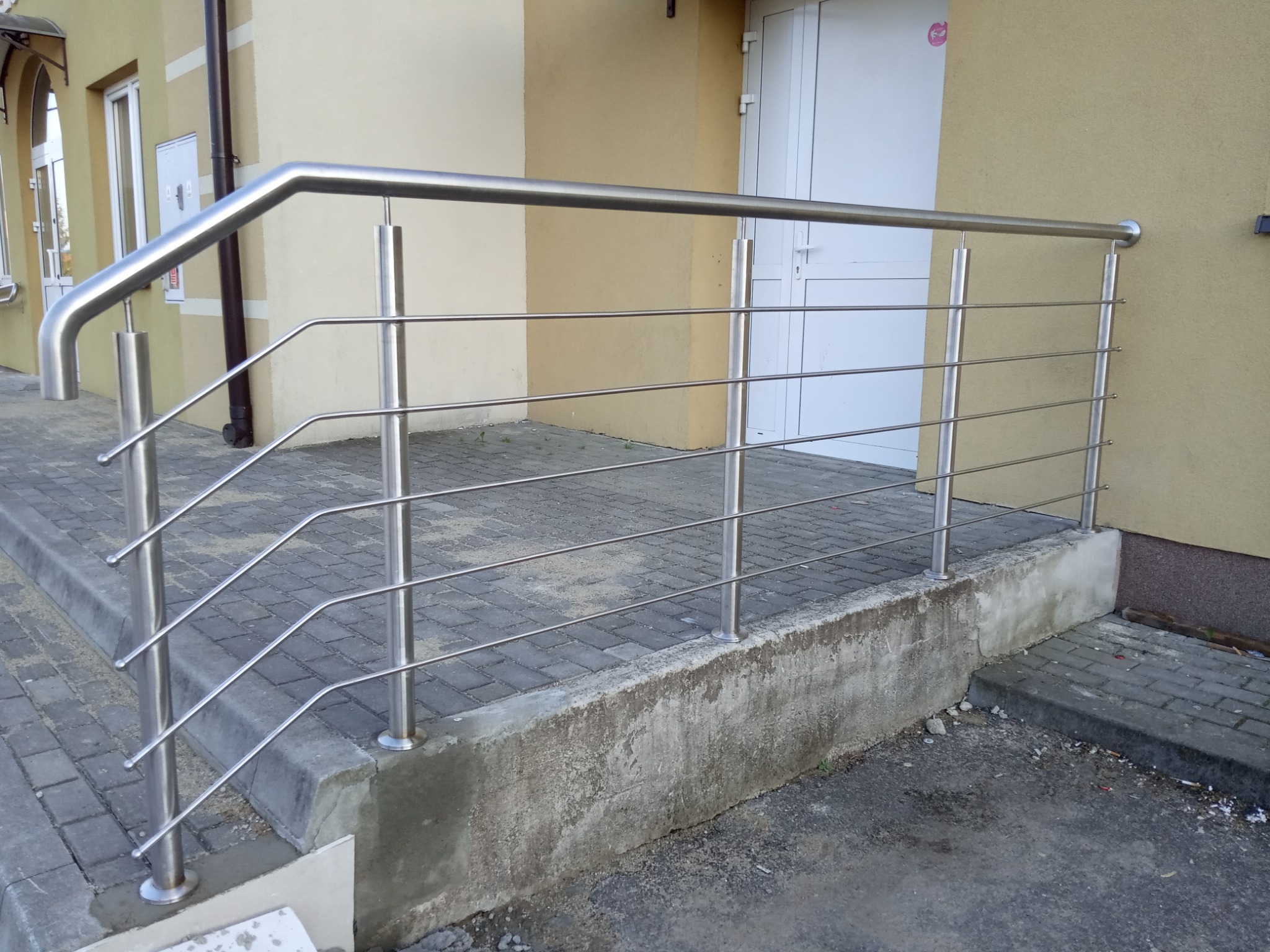 Zewnętrzna balustrada schodowa ze stali nierdzewnej, z czterema poziomymi prętami, zamocowana do betonowego podestu i ściany budynku w kolorze beżowym.