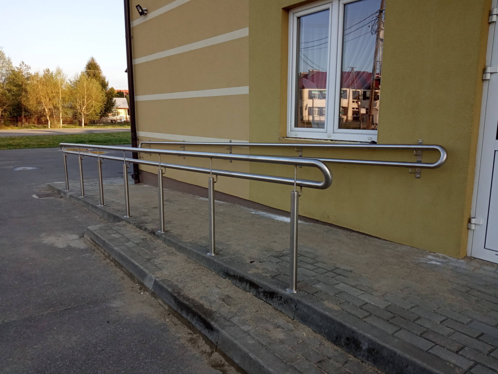 Zewnętrzna balustrada ze stali nierdzewnej przy wejściu do budynku, ułatwiająca dostęp dla osób z niepełnosprawnościami, z widocznym oknem i odbiciem budynku naprzeciwko.