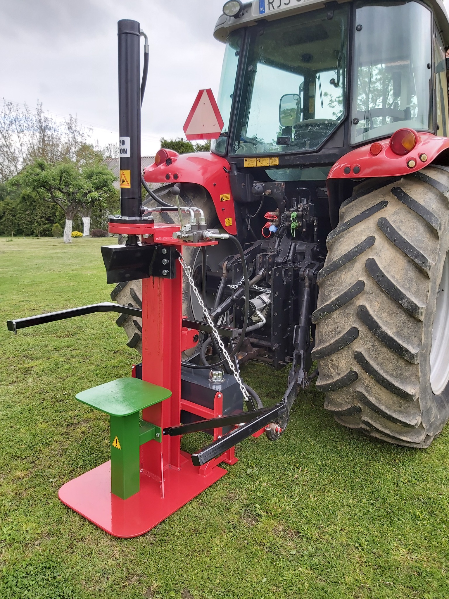 Czerwono-czarna łuparka do drewna z zielonym stołem roboczym, podłączona do czerwonego ciągnika rolniczego na trawniku.