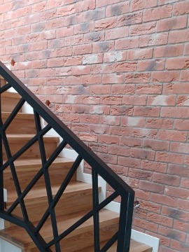 Klatka schodowa z drewnianymi stopniami, czarną metalową balustradą o geometrycznym wzorze i ścianą wyłożoną cegłą klinkierową w odcieniach czerwieni i brązu. Widoczne fragmenty białej listwy...