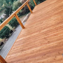 Nowoczesny taras z drewna o ciepłym odcieniu, z balustradą z drewnianych słupków i stalowych linek, widok z góry na deski i otoczenie z zielenią oraz budynkami w tle.