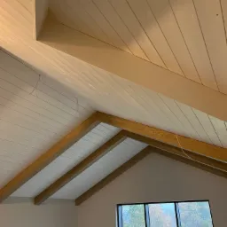 Wnętrze z drewnianym, skośnym sufitem wykończonym białą boazerią i bejcowanymi belkami, widok na jesienny krajobraz przez duże okno.