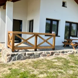Drewniana balustrada tarasowa z krzyżowym wzorem, zamontowana na kamiennej podmurówce przed białym domem z ciemnymi oknami, obok drewniany stół piknikowy.