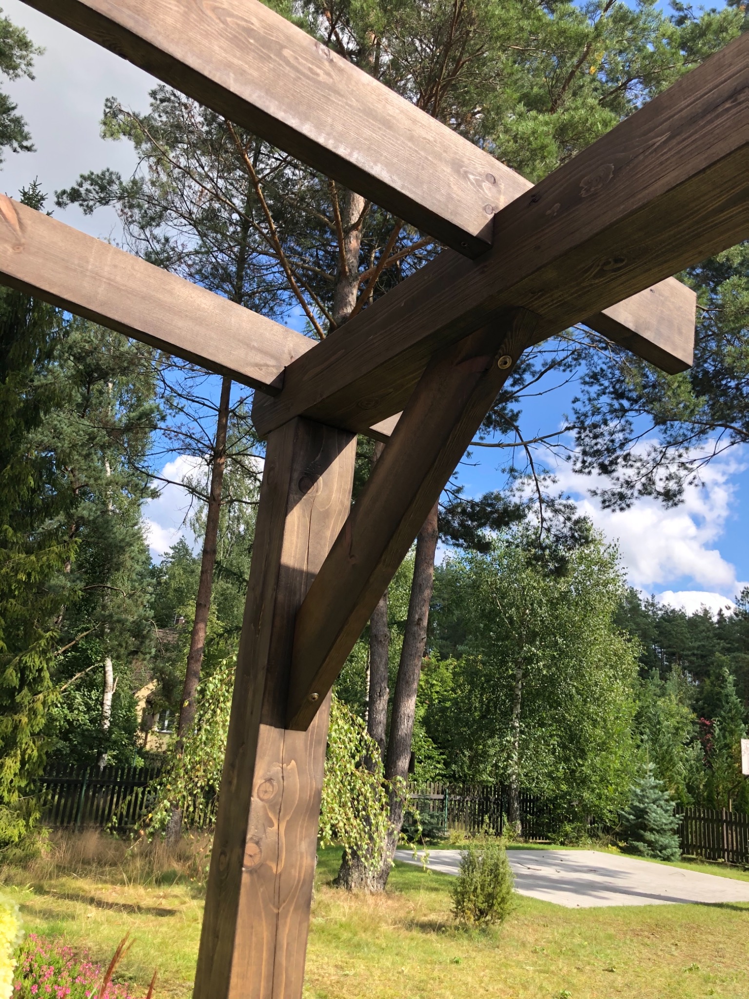 Fragment konstrukcji drewnianej, prawdopodobnie pergoli, wykonanej z ciemnego drewna, z widocznymi elementami mocującymi, na tle ogrodu z drzewami i błękitnym niebem z chmurami.
