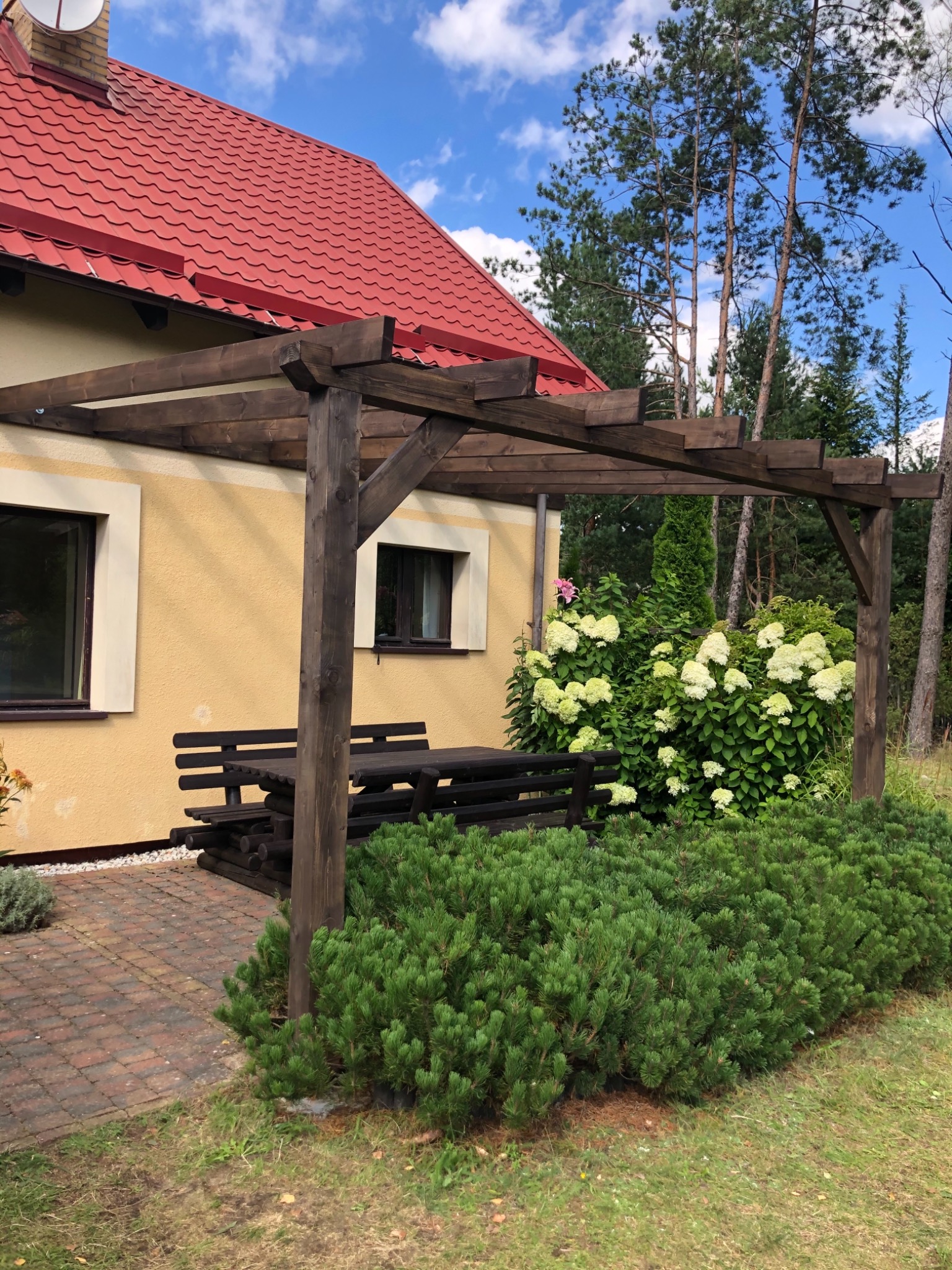 Ciemnobrązowa drewniana konstrukcja z ławką i stołem, przylegająca do ściany domu z czerwoną dachówką, otoczona zielenią, w tle drzewa iglaste i błękitne niebo z chmurami.