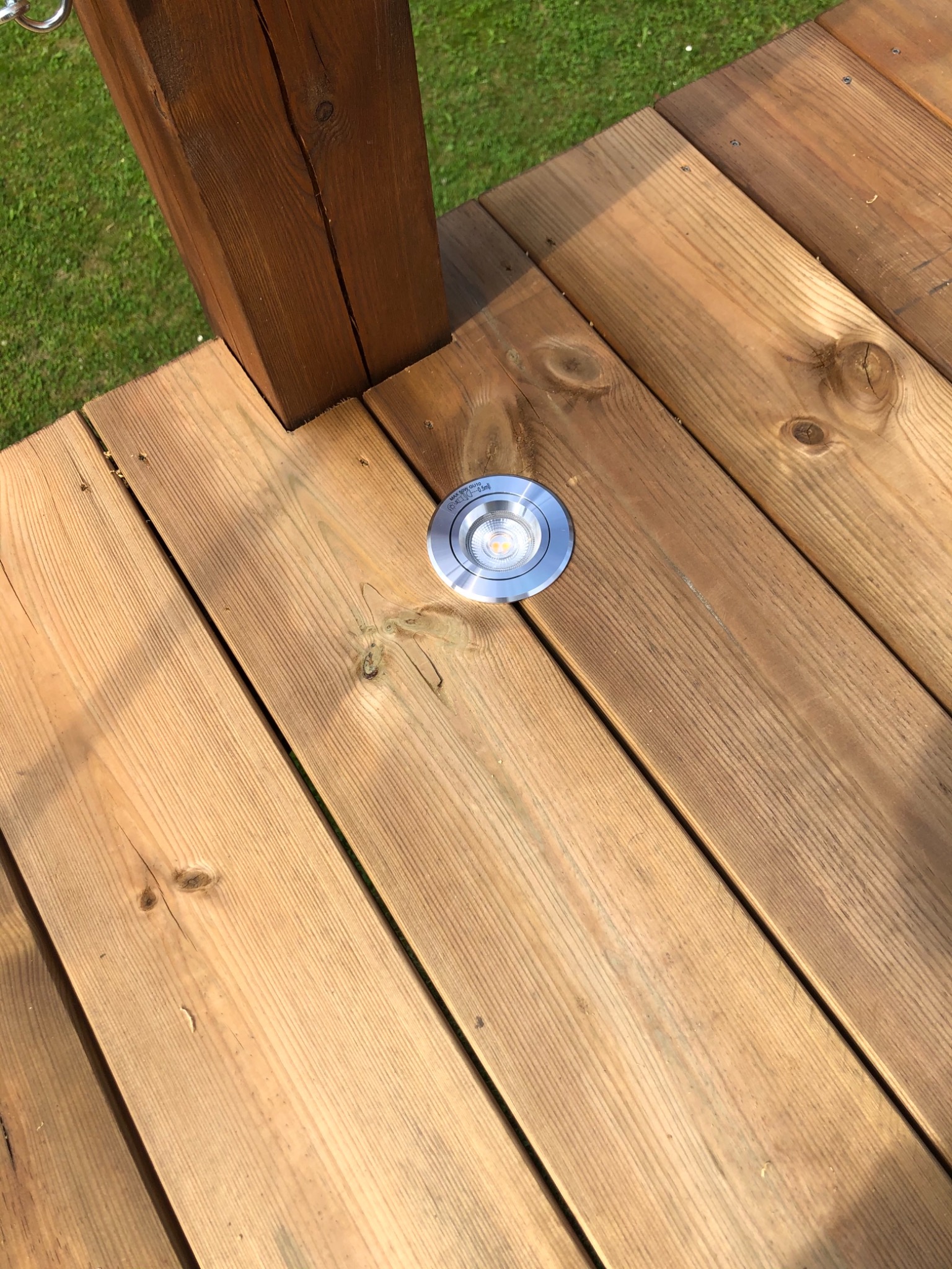 Fragment drewnianego tarasu z wbudowanym okrągłym oświetleniem LED, ukazujący naturalne usłojenie drewna i mocowanie do słupka konstrukcyjnego.