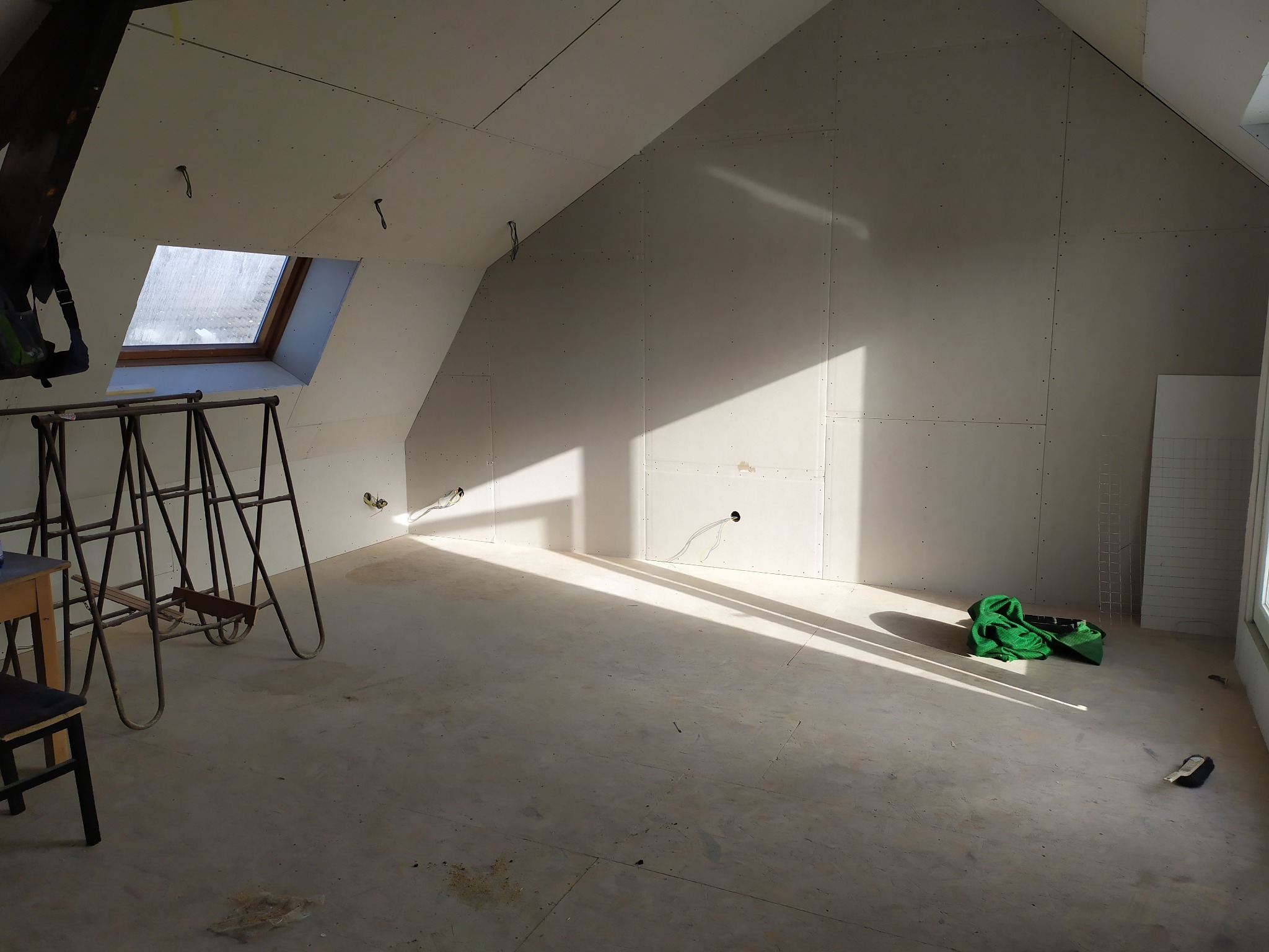 Poddasze w trakcie wykańczania, ściany pokryte płytami gipsowo-kartonowymi, widoczne narzędzia i drabina, wpadające promienie słoneczne podkreślają etap prac.