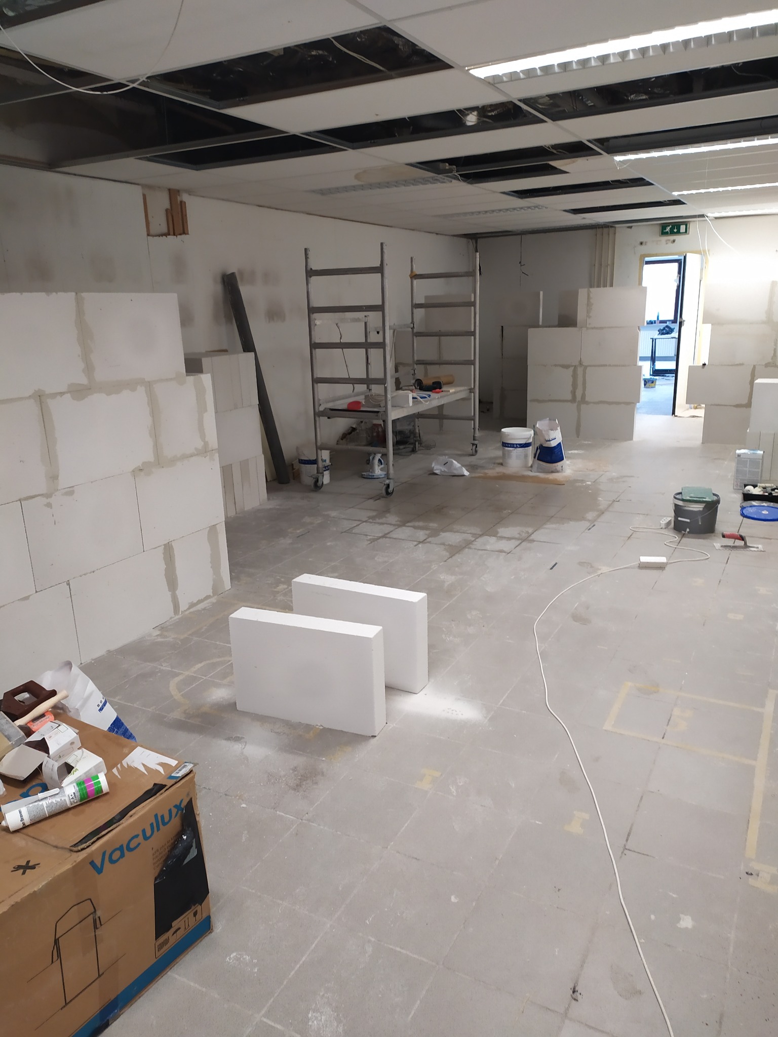 Wnętrze biura w trakcie prac budowlanych z widocznymi pustakami, rusztowaniem, workami z zaprawą i otwartym sufitem podwieszanym.