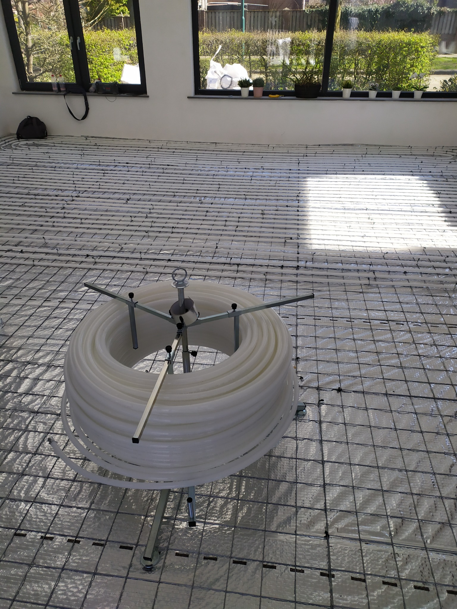 Rozłożona instalacja ogrzewania podłogowego z rurami na metalowej siatce, widoczny zwój rur na stojaku, duże okna w tle.