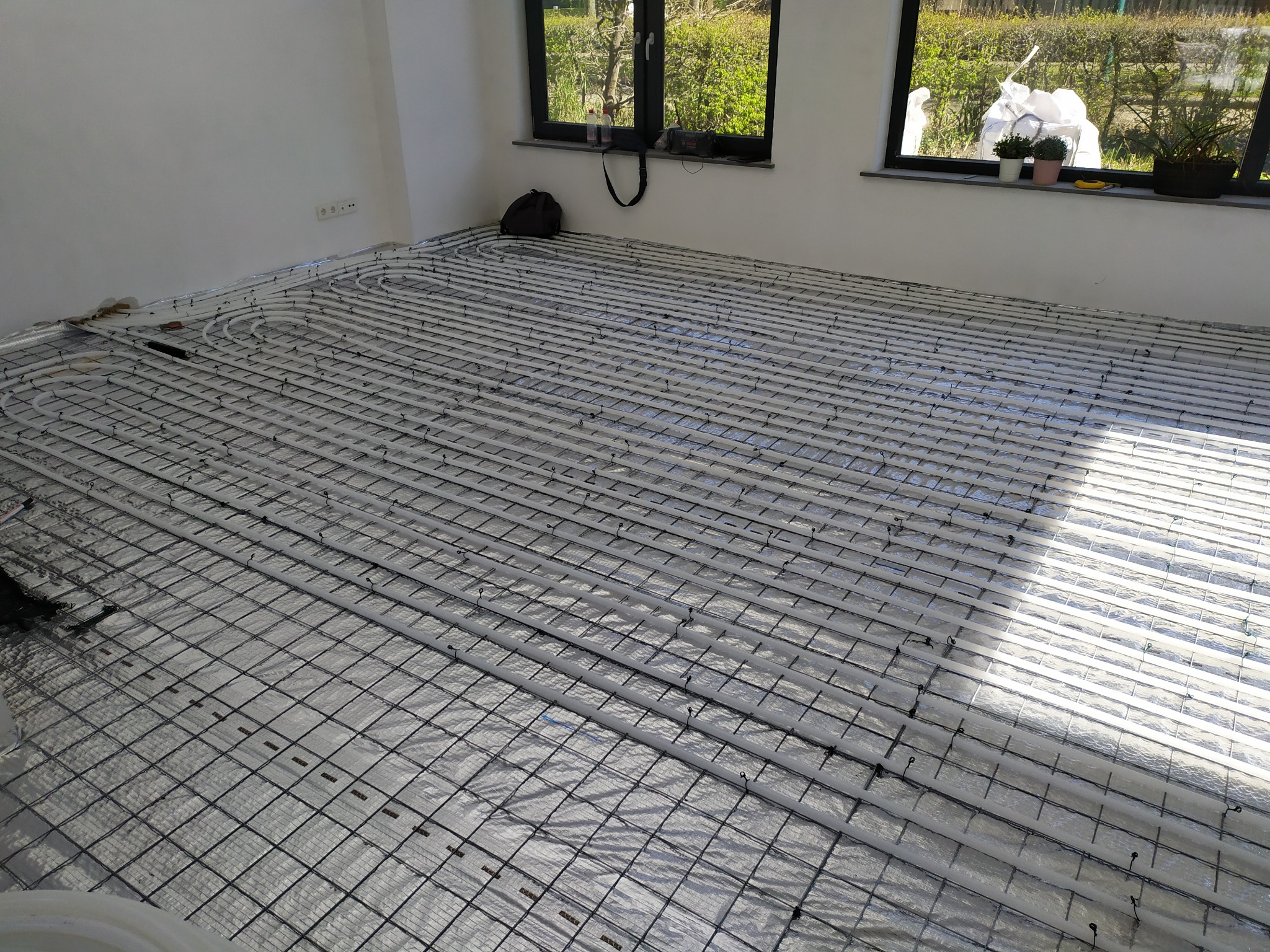 Montaż ogrzewania podłogowego: białe rurki ułożone w regularnych odstępach na folii odblaskowej z metalową siatką, widok na jasne wnętrze z oknami.