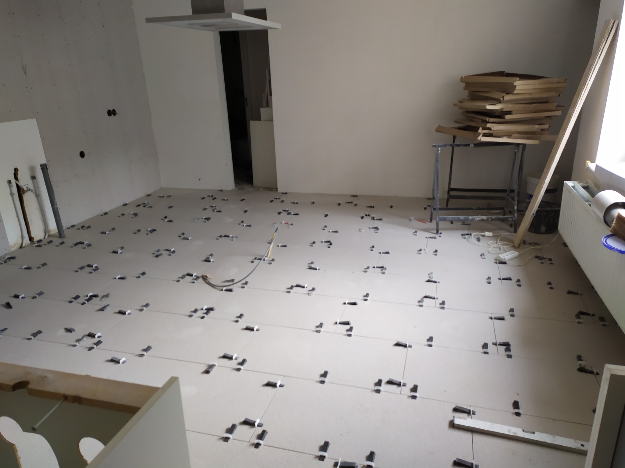 Białe płytki podłogowe w trakcie układania, widoczne czarne klipsy poziomujące i przewód elektryczny na podłodze, w tle stół z ułożonymi płytkami i grzejnik.