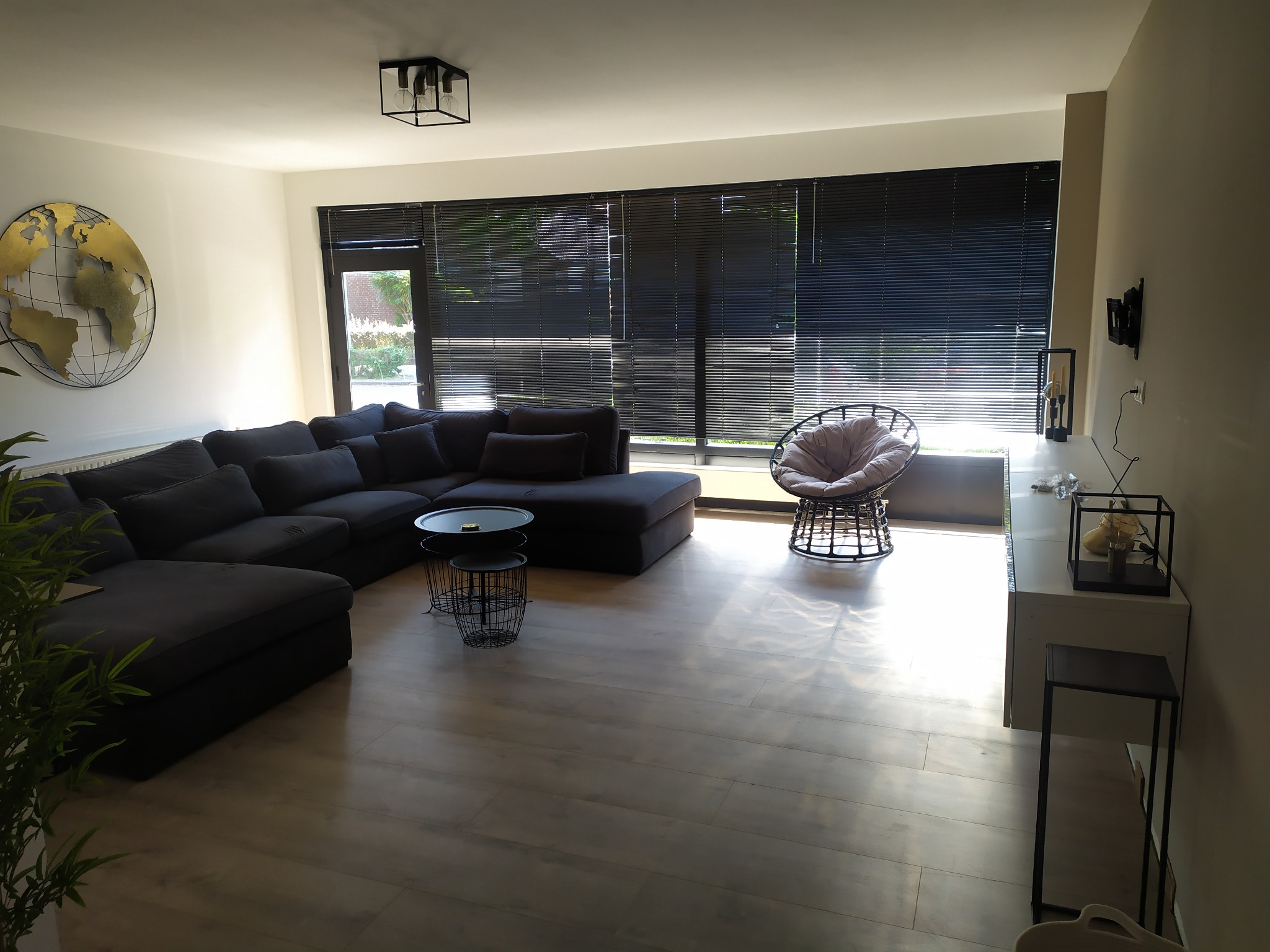 Jasne wnętrze salonu z szarym narożnikiem, drewnianą podłogą i czarnymi żaluzjami w oknach, oświetlone popołudniowym słońcem.