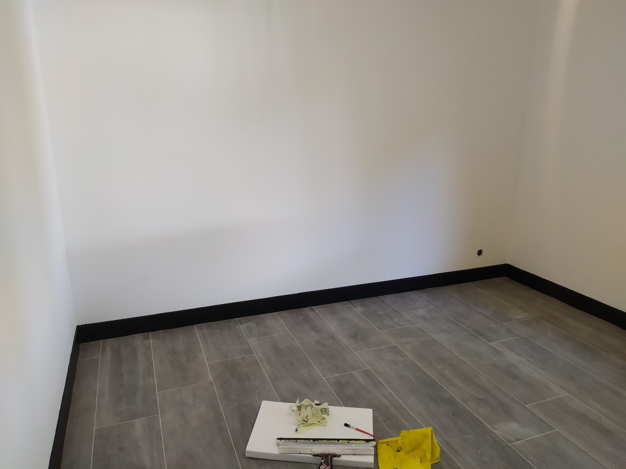 Pokój po remoncie z szarymi panelami podłogowymi imitującymi drewno, czarnymi listwami przypodłogowymi i białymi ścianami. Na podłodze leżą narzędzia: szpachelka, ściereczka i wiadro.