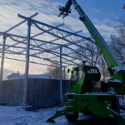 Montaż stalowej konstrukcji hali z użyciem zielonej ładowarki teleskopowej w zimowej scenerii, widoczne ośnieżone drzewa i częściowo betonowy fundament.