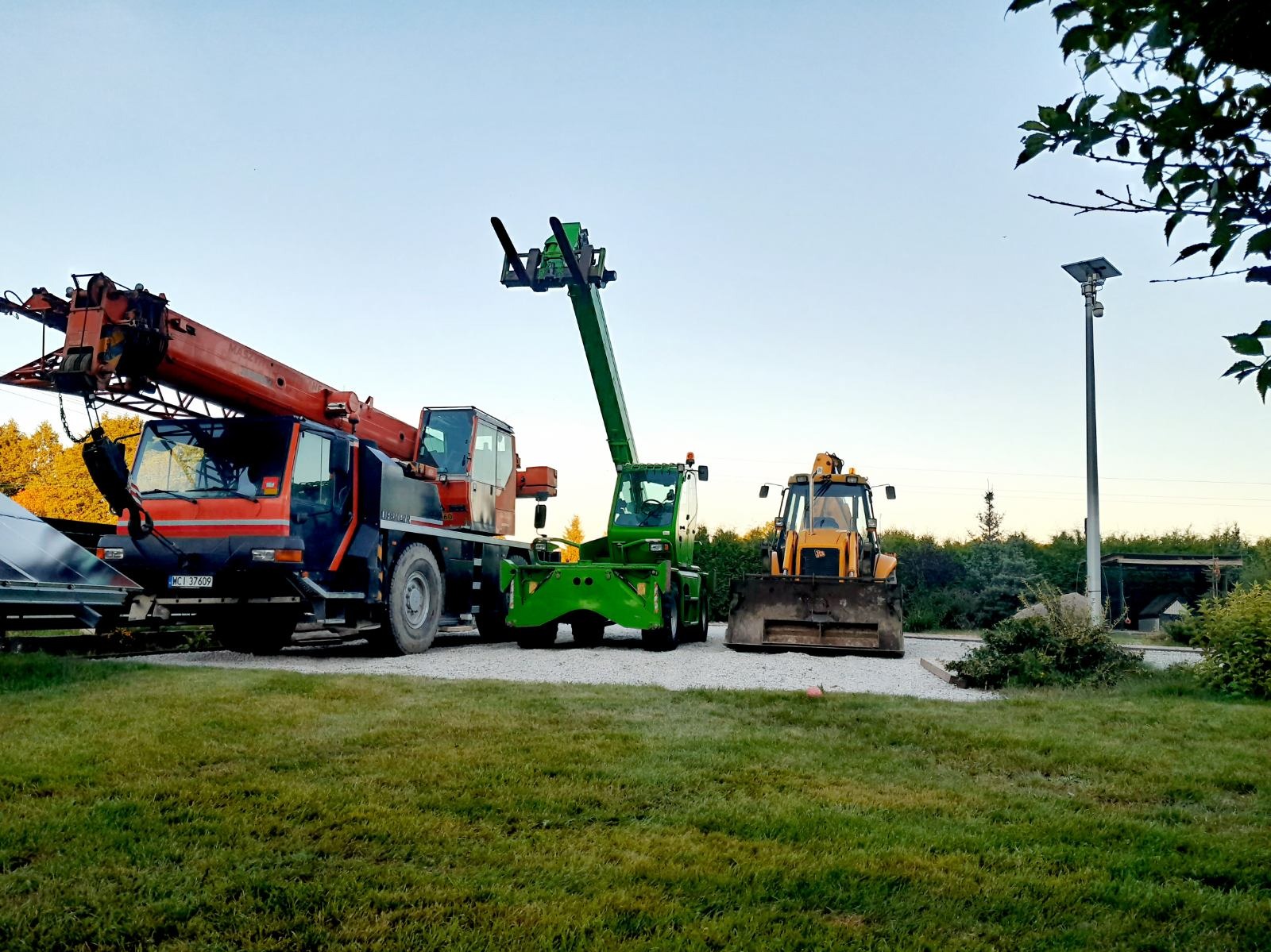 Ciężki sprzęt budowlany: pomarańczowy dźwig samochodowy Liebherr, zielony wózek teleskopowy i żółta koparko-ładowarka JCB, stojące na żwirowym podłożu obok trawnika w świetle wczesnego poranka.