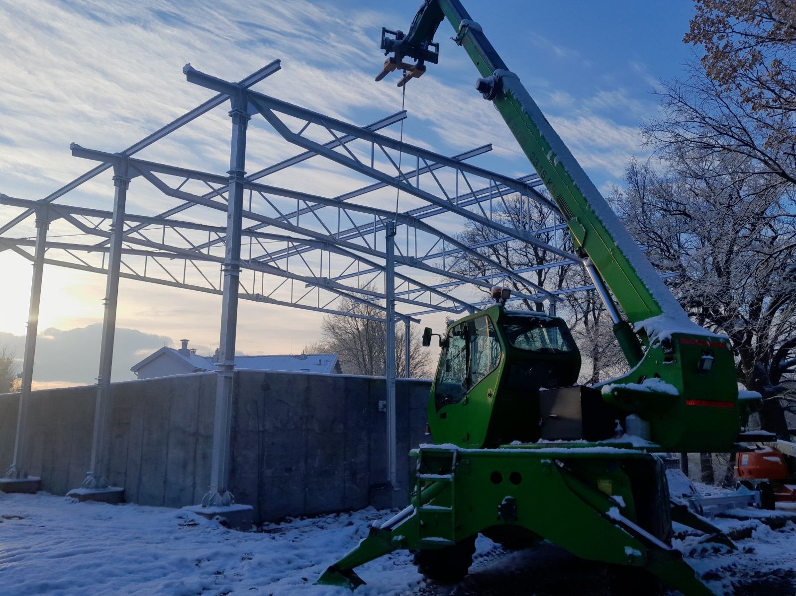 Montaż stalowej konstrukcji hali z użyciem zielonej ładowarki teleskopowej w zimowej scenerii, widoczne ośnieżone drzewa i częściowo betonowy fundament.