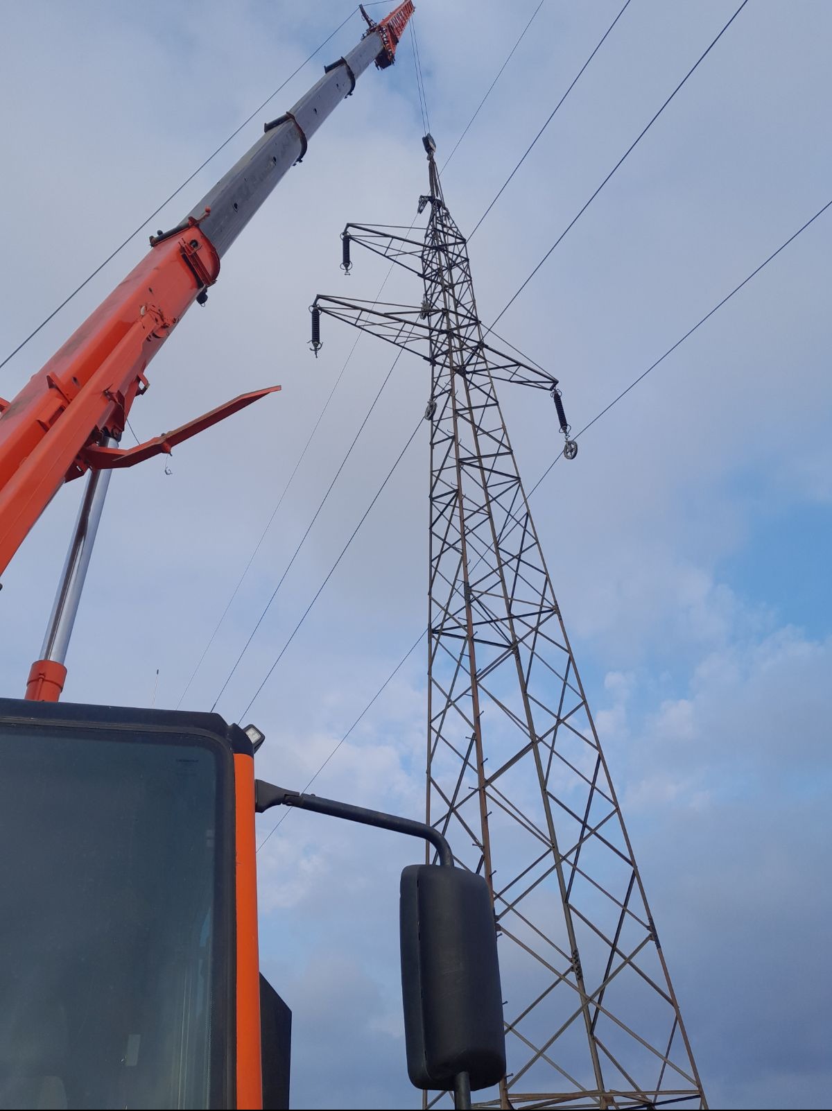 Pomarańczowy dźwig teleskopowy podnosi element konstrukcyjny stalowej wieży energetycznej na tle błękitnego nieba z chmurami.