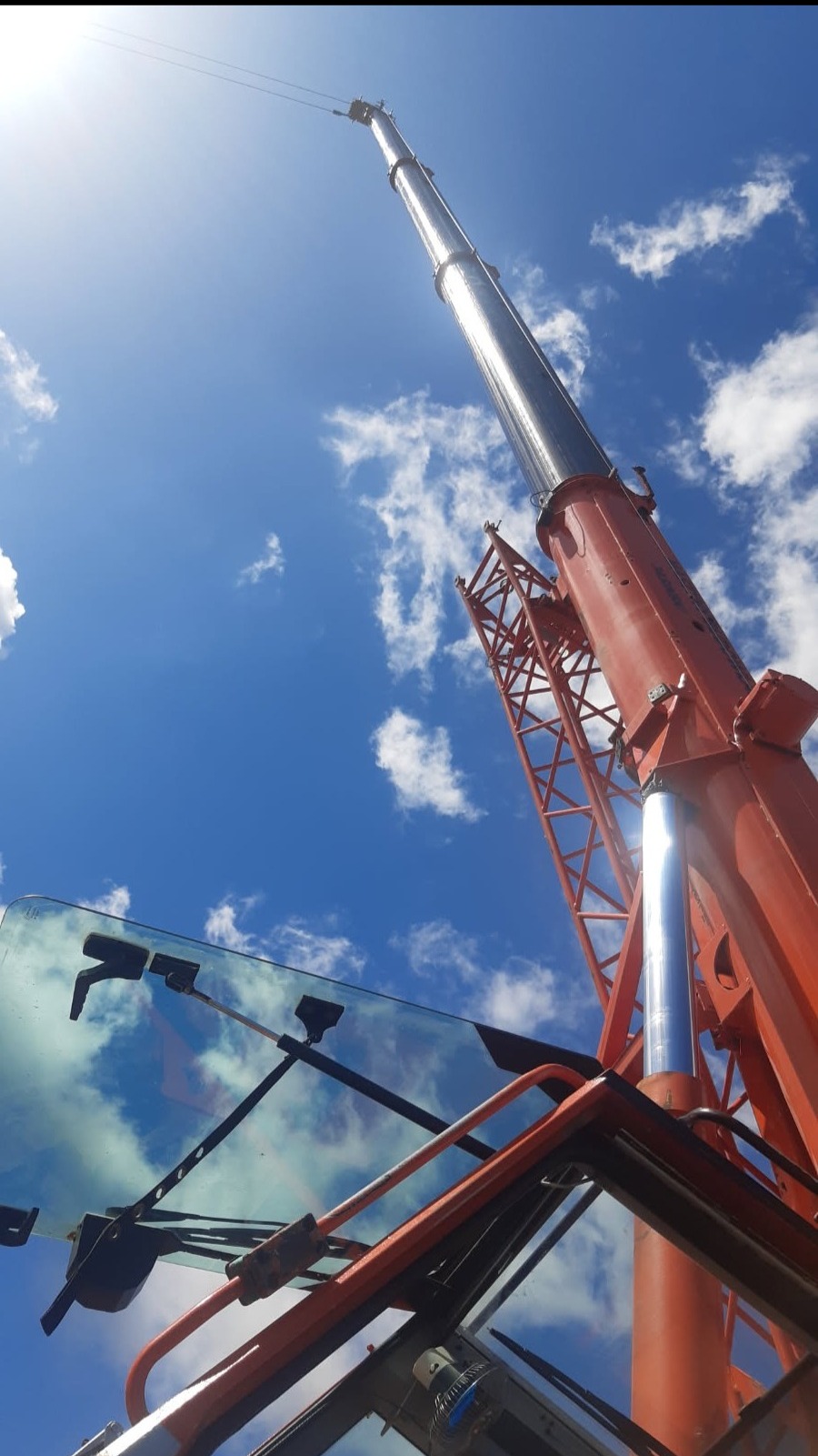Widok z kabiny na wysięgnik pomarańczowego dźwigu teleskopowego, skierowany w górę, na tle błękitnego nieba z chmurami.