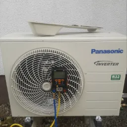 Biała jednostka zewnętrzna klimatyzacji Panasonic Inverter R32, podłączona do cyfrowego manometru Testo 550 za pomocą żółtego i niebieskiego węża, ustawiona na betonowych wspornikach na żwirowym...