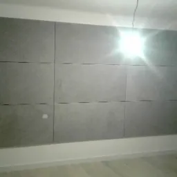 Ściana wykończona panelami z płyty kartonowo-gipsowej w szarym odcieniu, widoczne łączenia paneli, fragment ściany z tapetą w pionowe pasy oraz krzesło w rogu pokoju. Oświetlenie sufitowe tworzy...