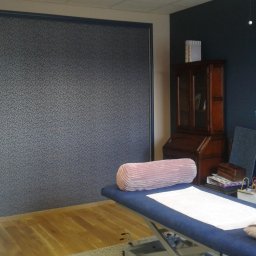 Dominik Hanek - Pokój z tapetą w drobny wzór, drewnianą podłogą i niebieskim łóżkiem rehabilitacyjnym z poduszkami, widok na białe drzwi i antyczny kredens.