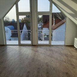 Dominik Hanek - Jasne poddasze z nową, brązową podłogą z płytek imitujących drewno, duże okna balkonowe z widokiem na zabudowę jednorodzinną, białe grzejniki.