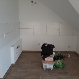 Dominik Hanek - Poddasze w trakcie prac wykończeniowych kuchni: ułożone płytki na ścianie, widoczne przyłącza wodne, na podłodze brązowe płytki imitujące drewno, kartony z płytkami i torba z narzędziami.