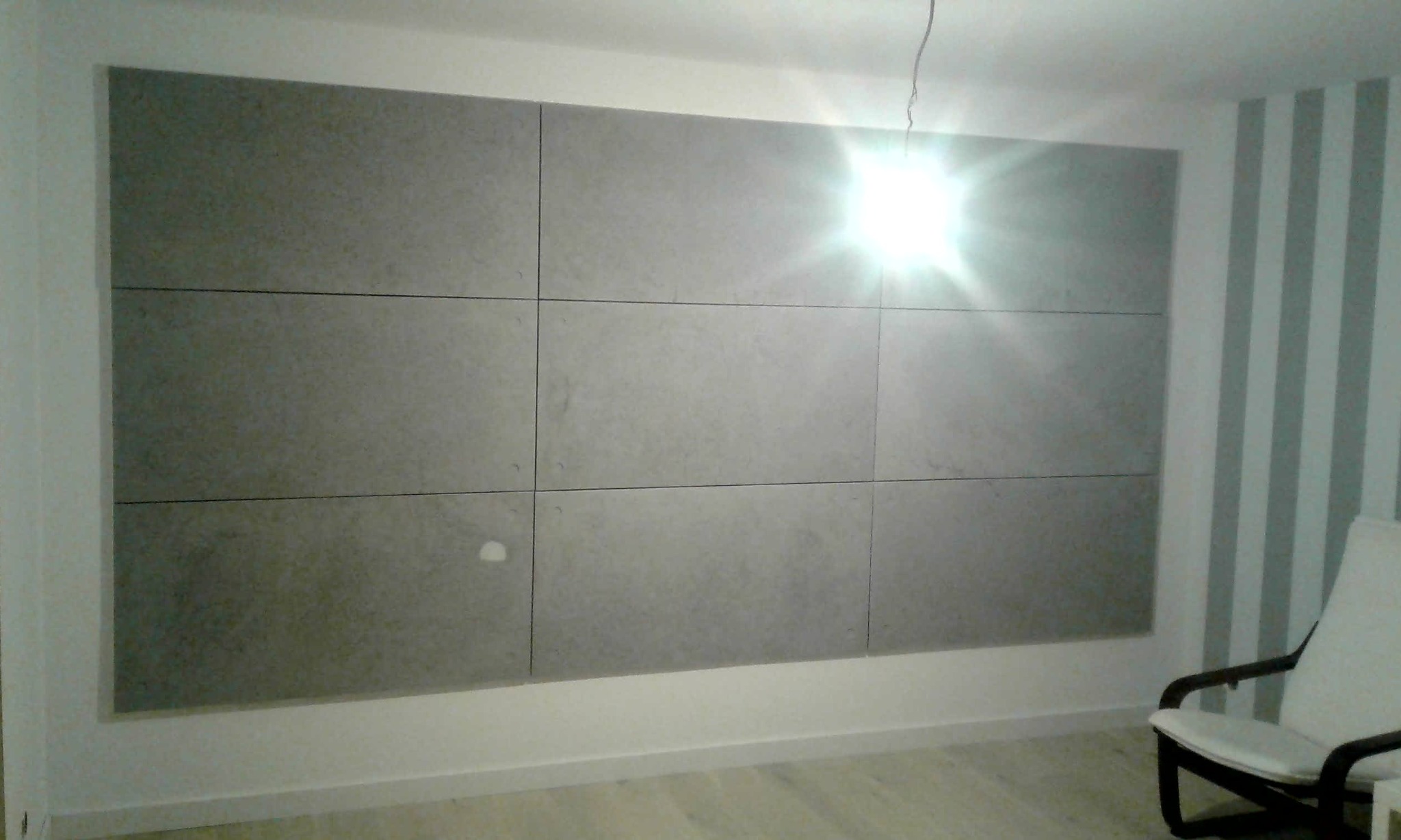 Ściana wykończona panelami z płyty kartonowo-gipsowej w szarym odcieniu, widoczne łączenia paneli, fragment ściany z tapetą w pionowe pasy oraz krzesło w rogu pokoju. Oświetlenie sufitowe tworzy...