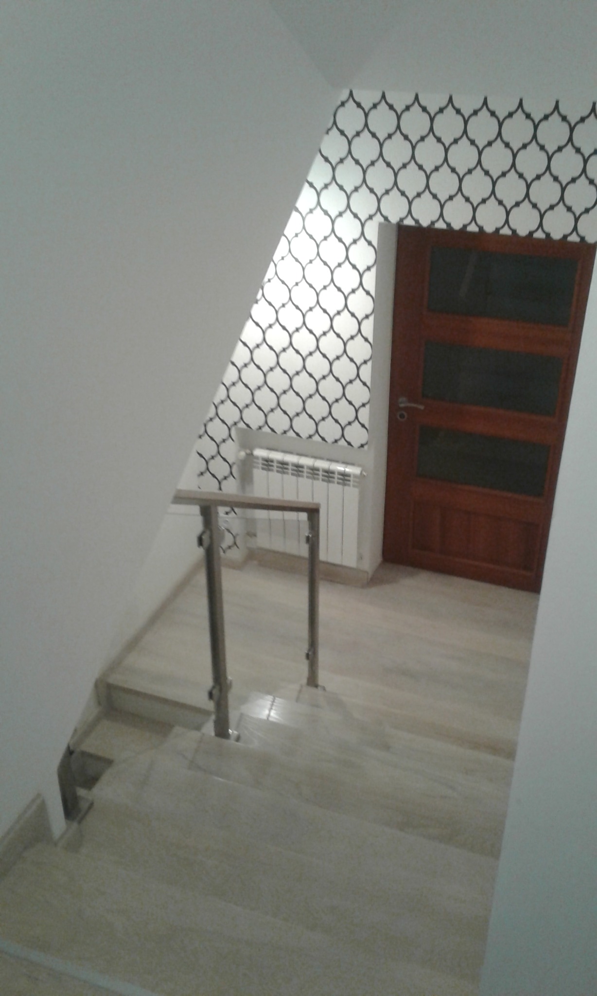 Klatka schodowa z marmurowymi schodami i metalową balustradą, ściana z tapetą w geometryczny wzór, drzwi z szybami i grzejnik.