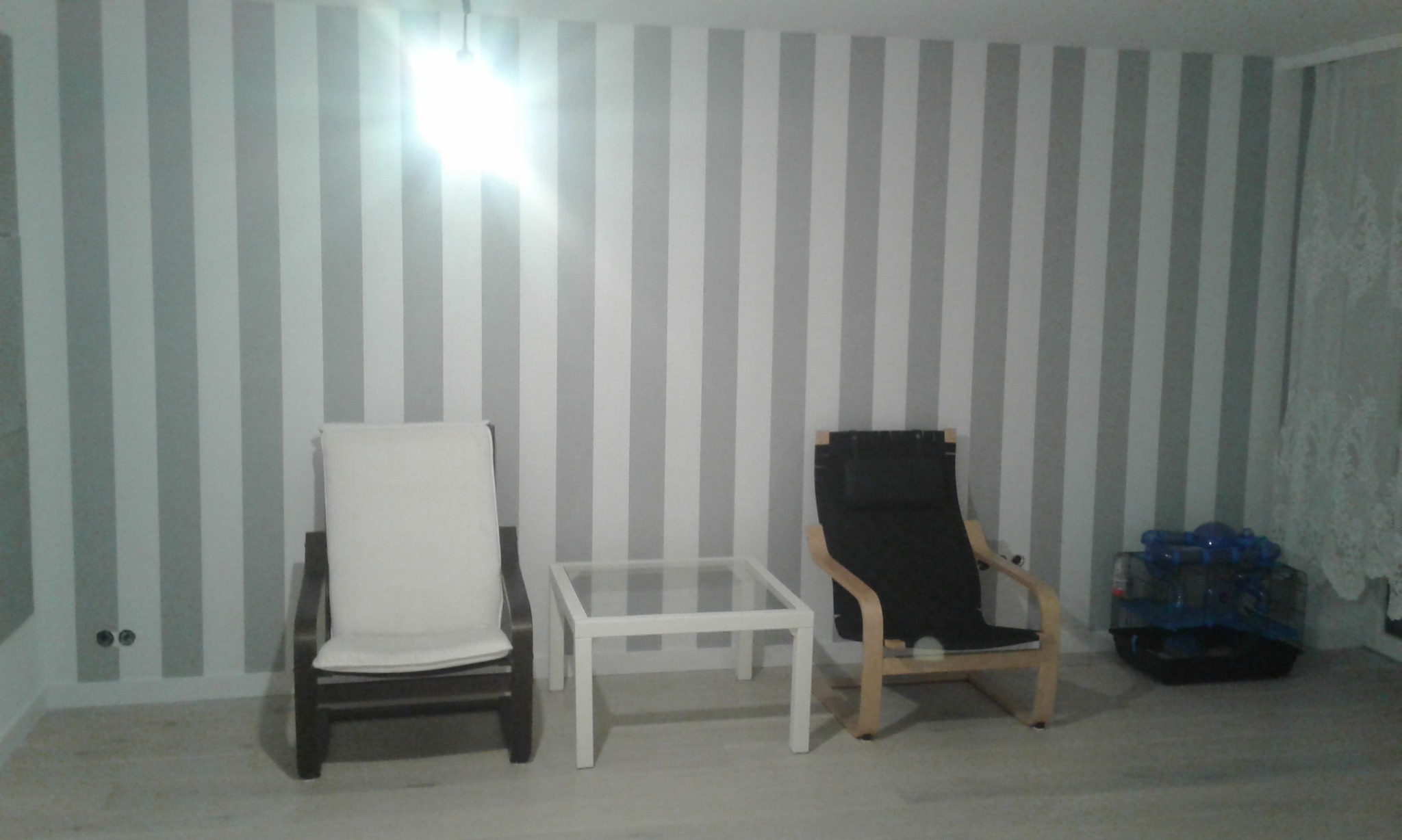 Pokój z tapetą w szaro-białe pasy, dwa fotele, szklany stolik i klatka dla zwierząt w rogu.