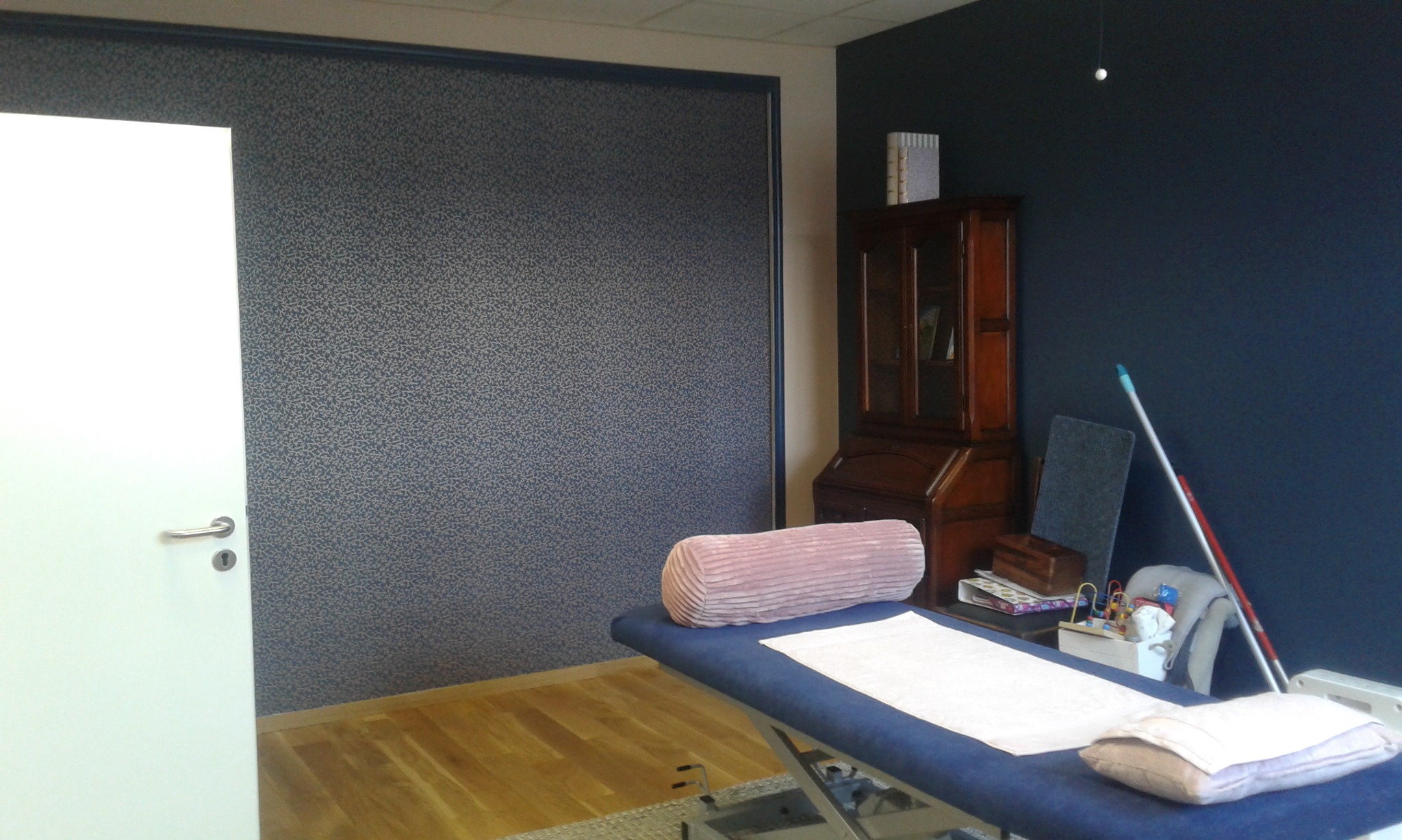 Pokój z tapetą w drobny wzór, drewnianą podłogą i niebieskim łóżkiem rehabilitacyjnym z poduszkami, widok na białe drzwi i antyczny kredens.