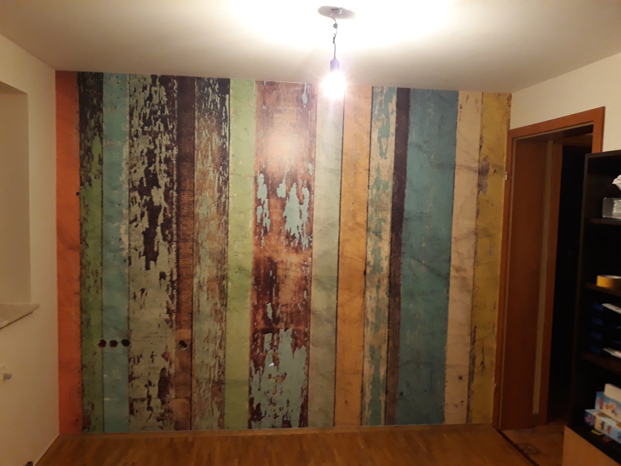 Ściana z fototapetą imitującą stare deski w różnych kolorach, widoczne gniazdka elektryczne, drewniana podłoga, oświetlenie górne.