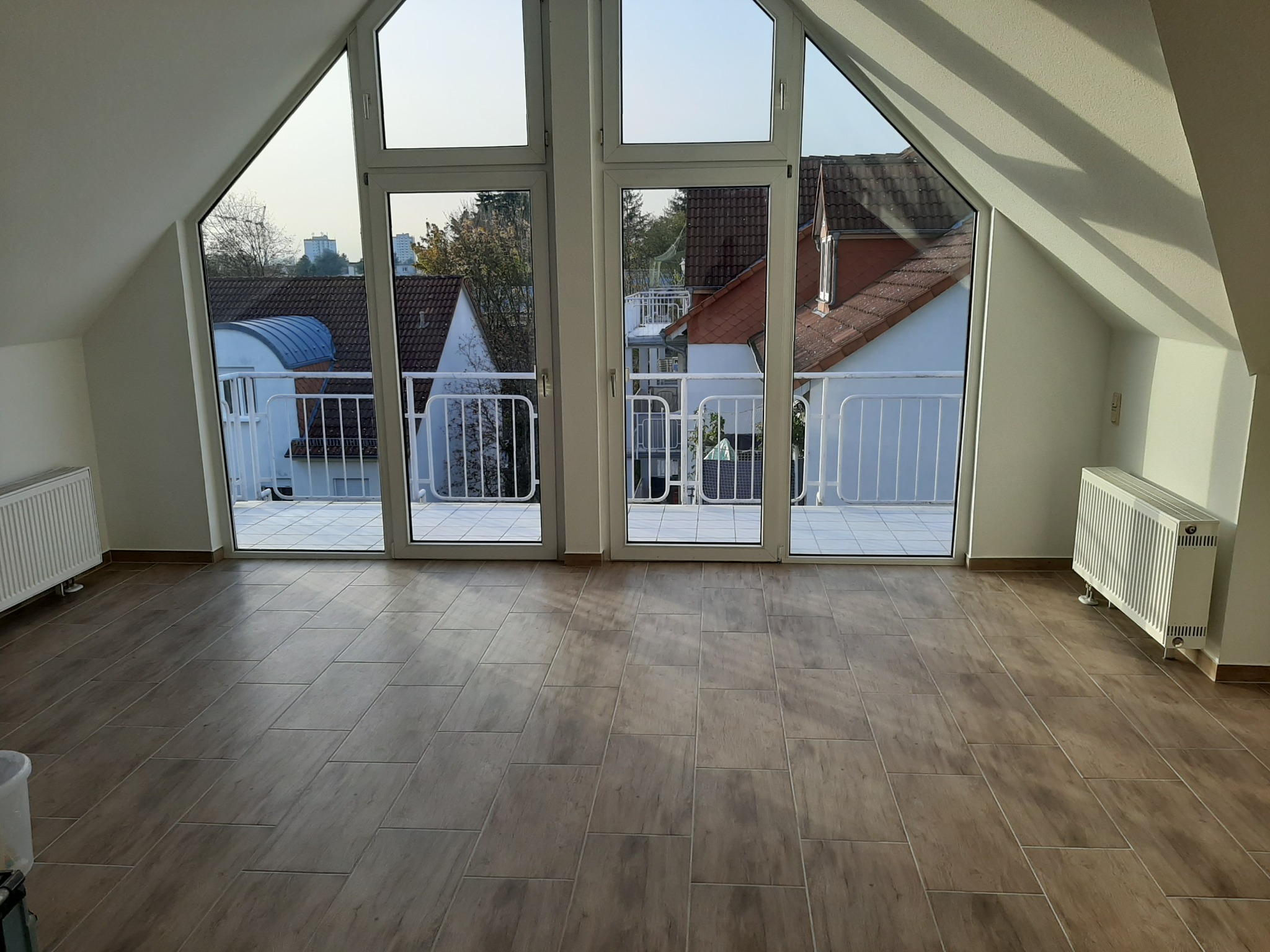 Jasne poddasze z nową, brązową podłogą z płytek imitujących drewno, duże okna balkonowe z widokiem na zabudowę jednorodzinną, białe grzejniki.