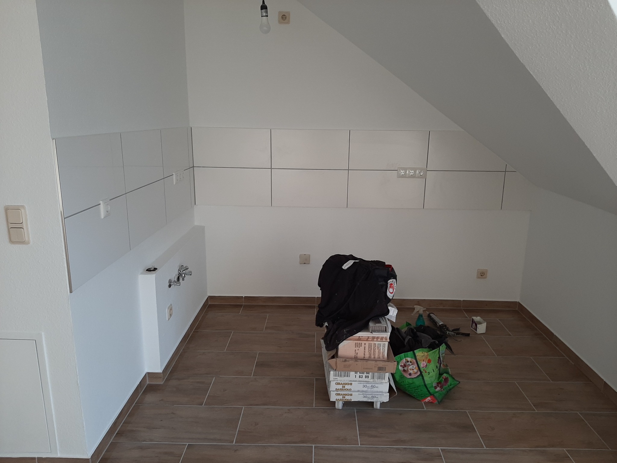 Poddasze w trakcie prac wykończeniowych kuchni: ułożone płytki na ścianie, widoczne przyłącza wodne, na podłodze brązowe płytki imitujące drewno, kartony z płytkami i torba z narzędziami.