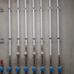 Rząd pionowych, białych rur z zaworami z niebieskimi pokrętłami, przymocowanych do szarej, betonowej ściany. Widoczne metalowe obejmy mocujące rury. Górna część rur biegnie poziomo pod sufitem.