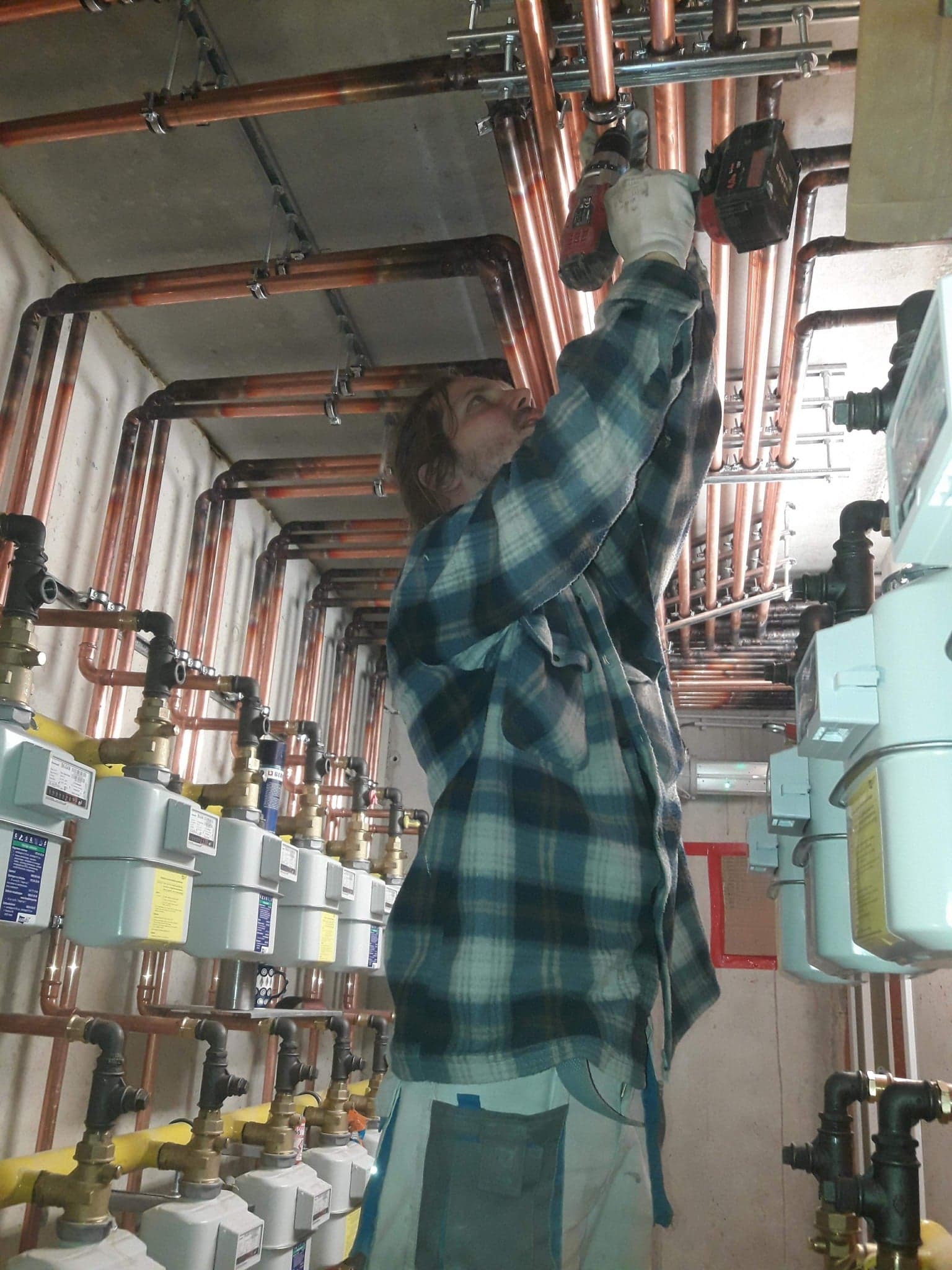 Instalator w koszuli w kratę wierci w miedzianej instalacji gazowej, widoczne liczniki gazu i rury pod sufitem.