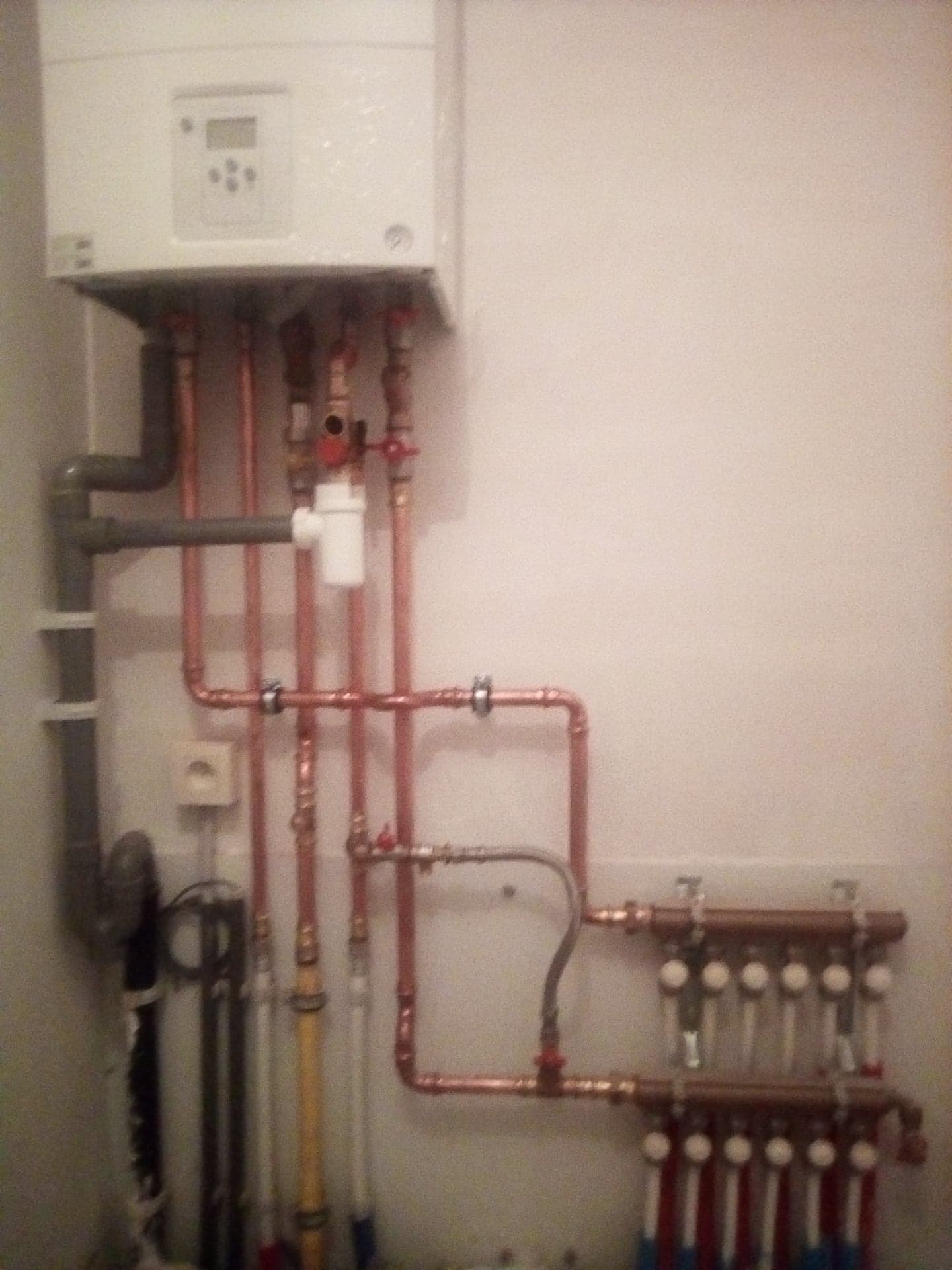 Biały piec gazowy z podłączonymi miedzianymi rurami instalacji centralnego ogrzewania i rozdzielaczem podłogowym z zaworami termostatycznymi, zamontowany na jasnej ścianie.