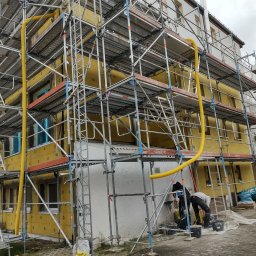 BUD-MIK MACIEJ MIKUNDA - Budynek w trakcie prac elewacyjnych, otoczony rusztowaniami. Widoczne żółte ocieplenie i pracownicy przygotowujący materiały. Charakterystyczna żółta rura wentylacyjna.