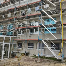 BUD-MIK MACIEJ MIKUNDA - Budynek w trakcie prac elewacyjnych, otoczony rusztowaniami. Widoczny pracownik w trakcie wykonywania prac na wysokości. Okna zabezpieczone folią.