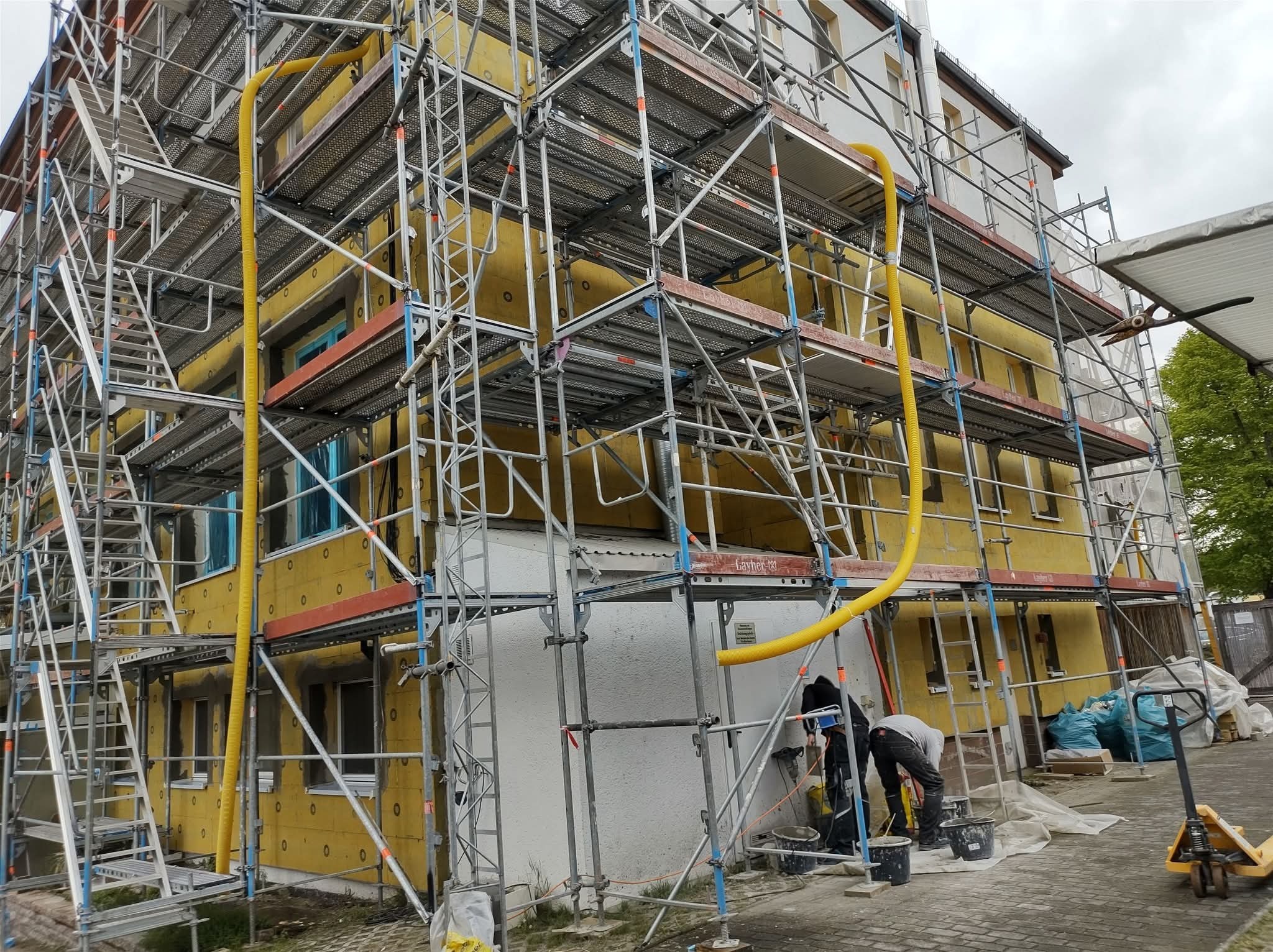 Budynek w trakcie prac elewacyjnych, otoczony rusztowaniami. Widoczne żółte ocieplenie i pracownicy przygotowujący materiały. Charakterystyczna żółta rura wentylacyjna.